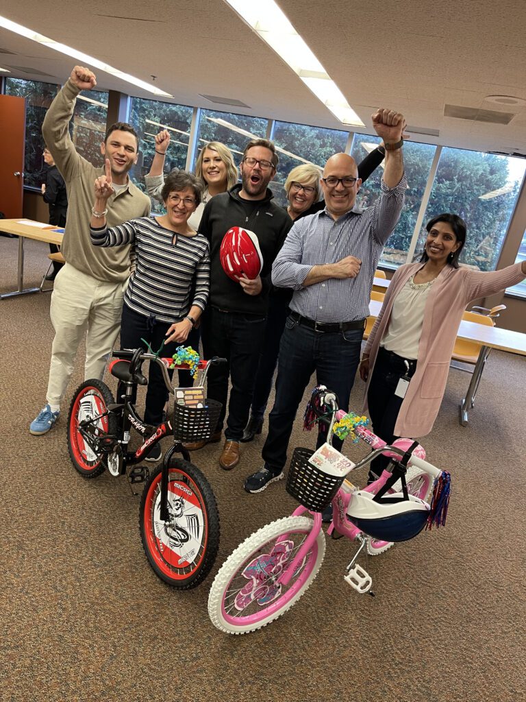 A group of seven participants cheer enthusiastically while standing behind two completed bikes during a Charity Bike Build event.