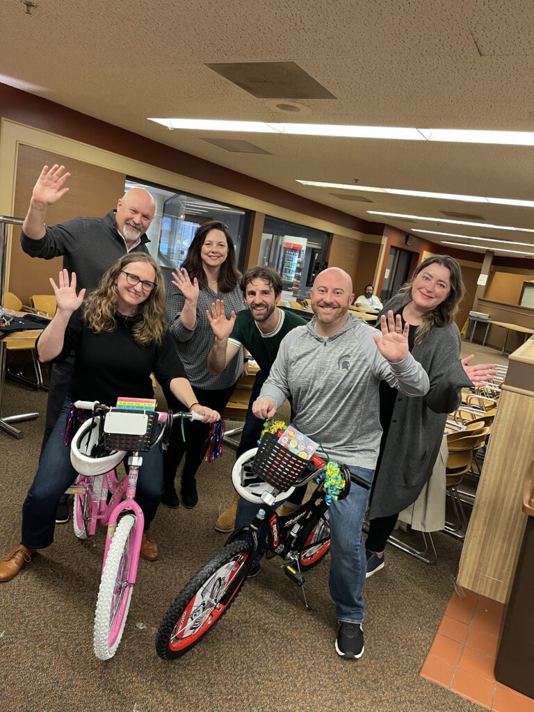 A group of six people smile and wave after successfully assembling two bicycles—one pink and one black—during a Charity Bike Build event. The team members stand proudly next to the bikes they built, showcasing their collaboration and effort.