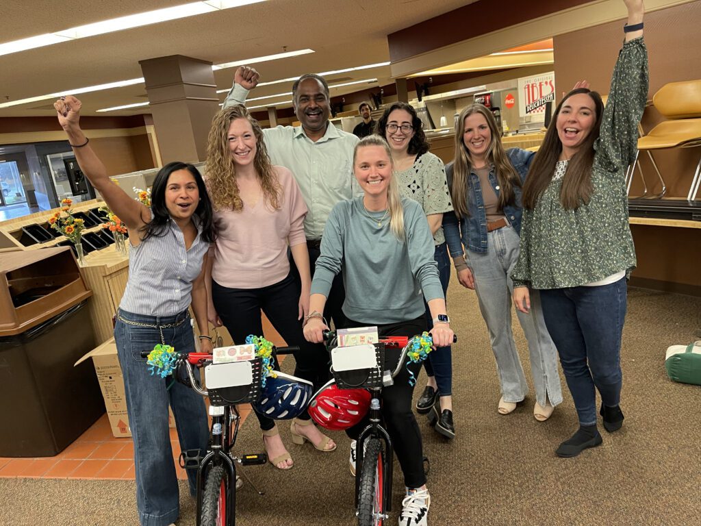 A cheerful group of seven team members celebrates their successful Charity Bike Build. Two decorated bikes stand in front of them, with the group raising their arms in victory. The atmosphere in the room is lively and filled with camaraderie.