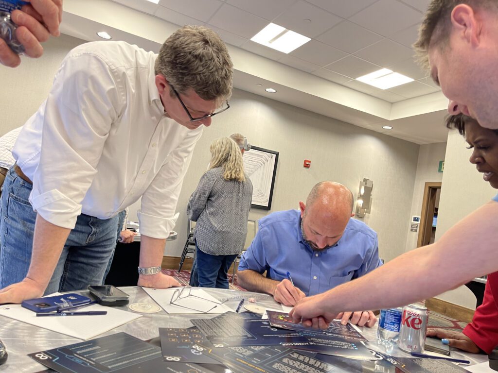 Participants gather around a table during The Infinite Loop team building event, focusing on solving a collaborative puzzle with strategy sheets laid out in front of them.