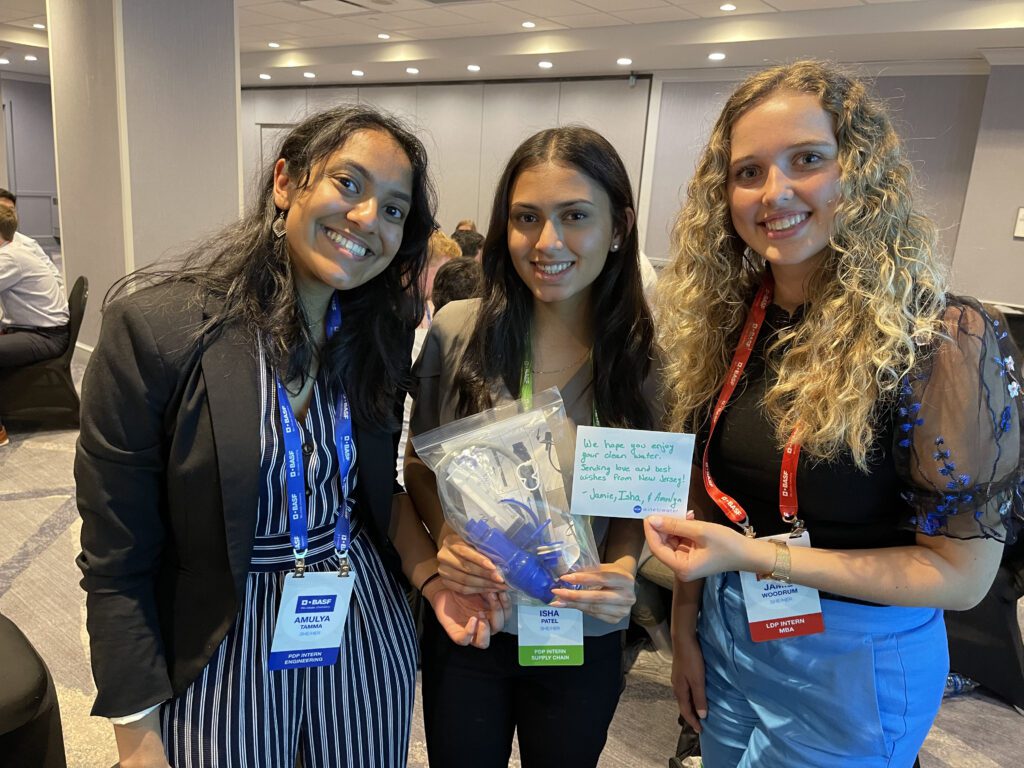 Three smiling women proudly display a completed water filtration kit and a handwritten note during a Clean Water Connection team building event.