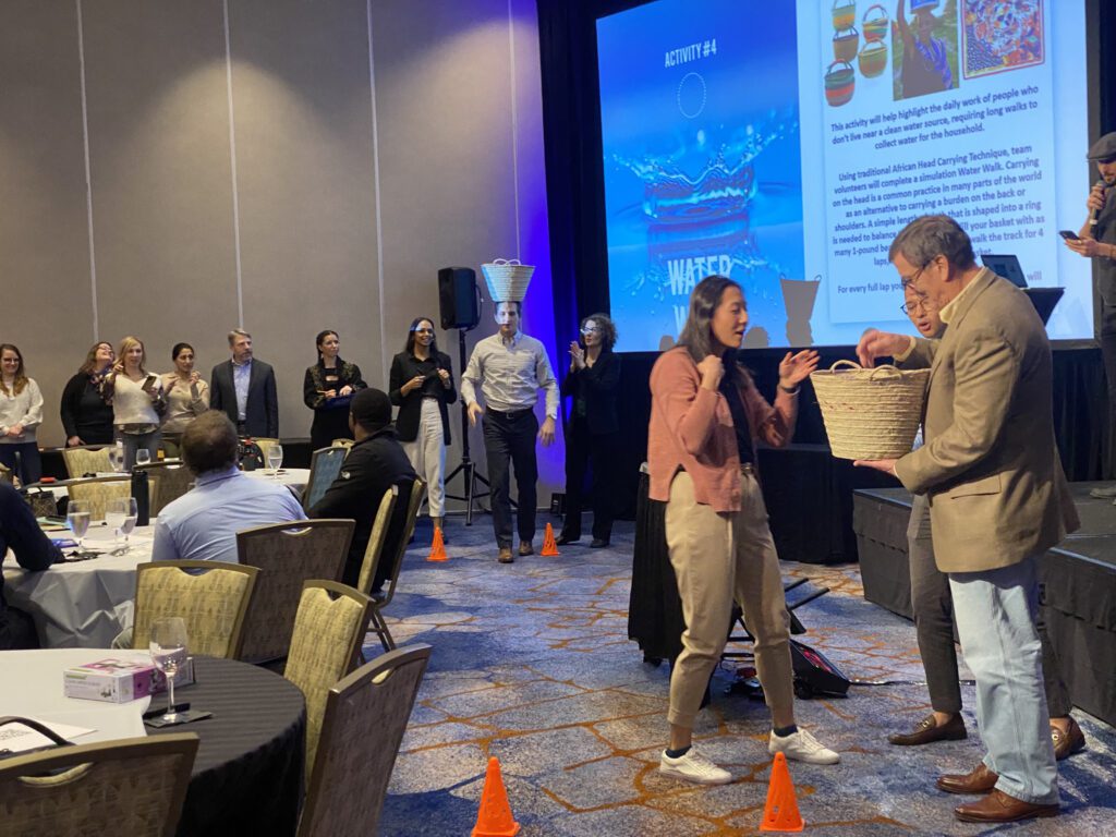 Participants engage in a water walk challenge during a Clean Water Connection team building event, balancing baskets to simulate the experience of collecting water in developing regions. The activity takes place in a conference setting, with a large screen in the background displaying information about water scarcity.