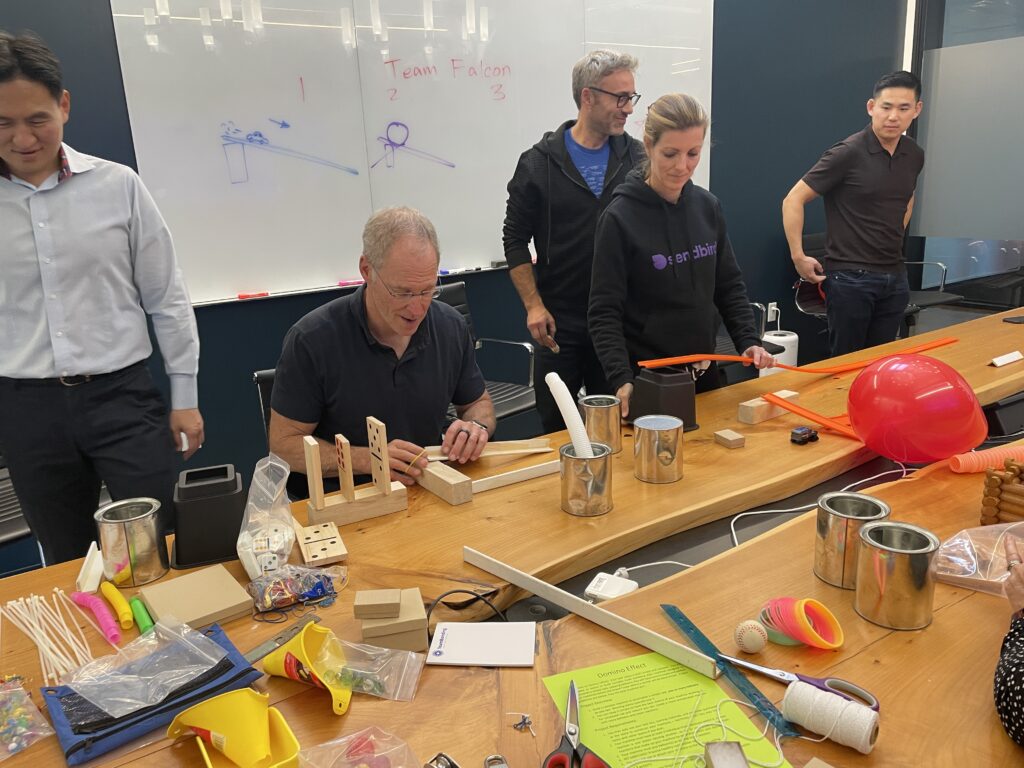 Team members collaborating and building creative contraptions during the Domino Effect team building event, surrounded by various building materials and tools.