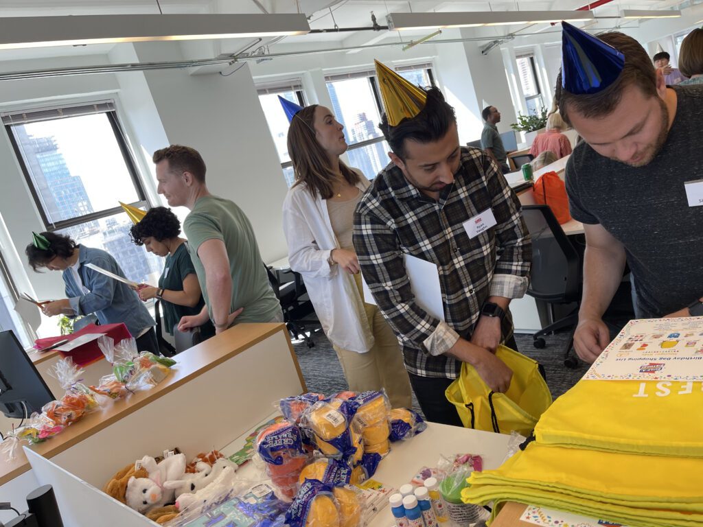 A group of people wearing colorful party hats are assembling gift bags at a long table filled with various supplies, such as stuffed animals, toys, and arts and crafts materials. Some individuals are focused on their tasks, while others are engaging in conversations. The scene reflects a collaborative and charitable team building event.