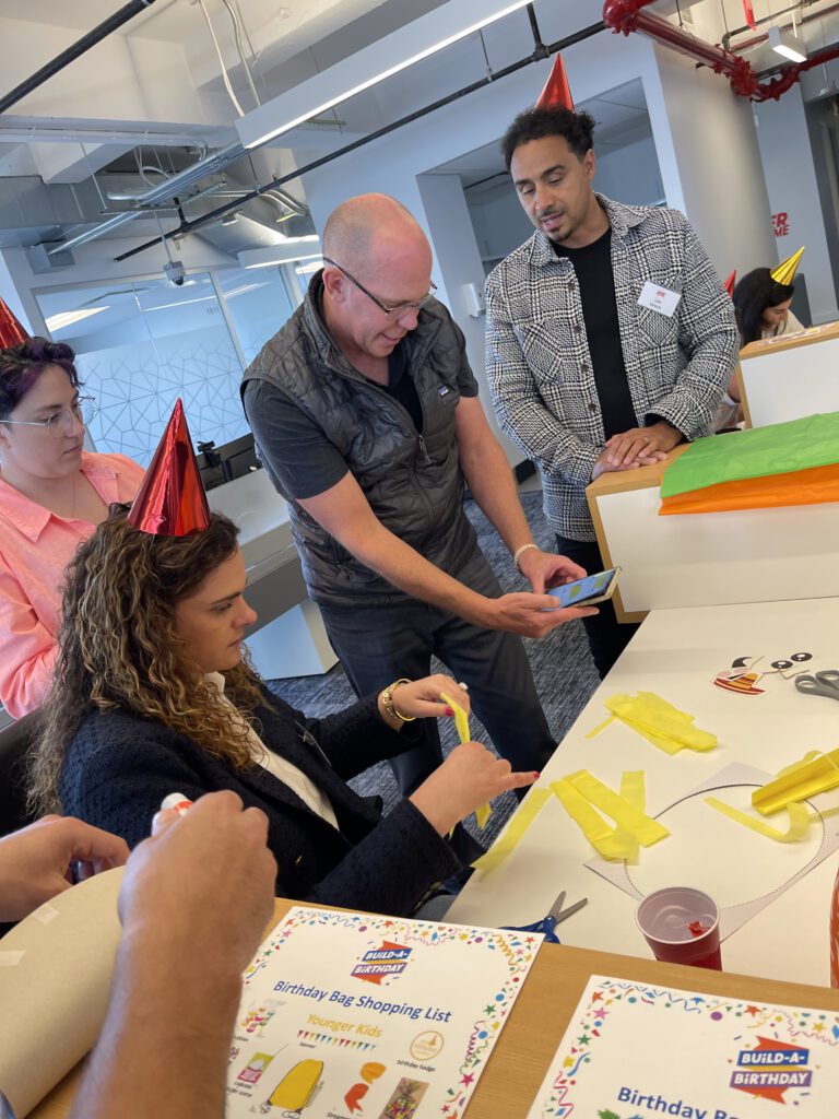 A group of people wearing party hats are engaged in a crafting activity. One person is seated, cutting yellow paper strips, while others stand around, observing and discussing the team building activity.