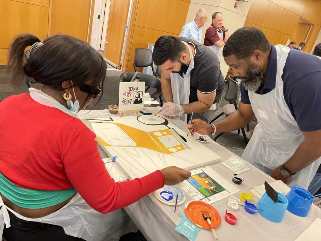 Team members collaborate to paint a section of a large mural during The Big Picture team building event, working together to create a unified masterpiece.