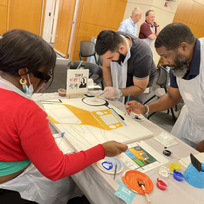 Team members collaborate to paint a section of a large mural during The Big Picture team building event, working together to create a unified masterpiece.