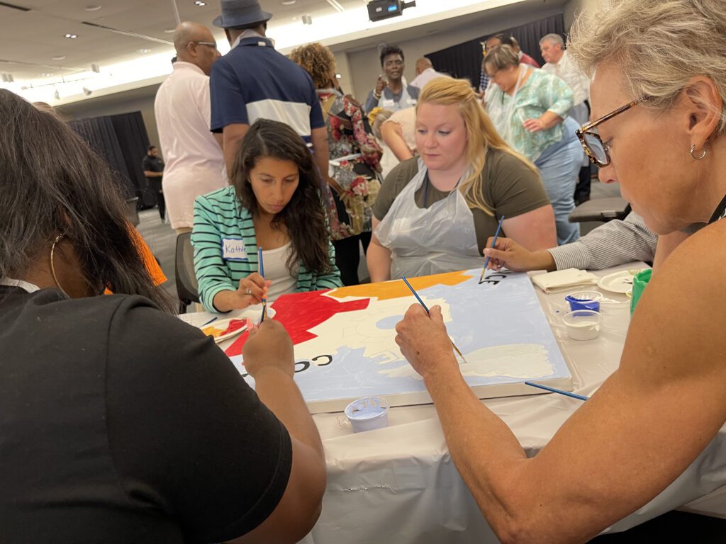 A group of participants sits around a table during The Big Picture team building event, painting a canvas section with various colors. They focus on filling in their assigned part of the mural, collaborating to complete the larger artwork.