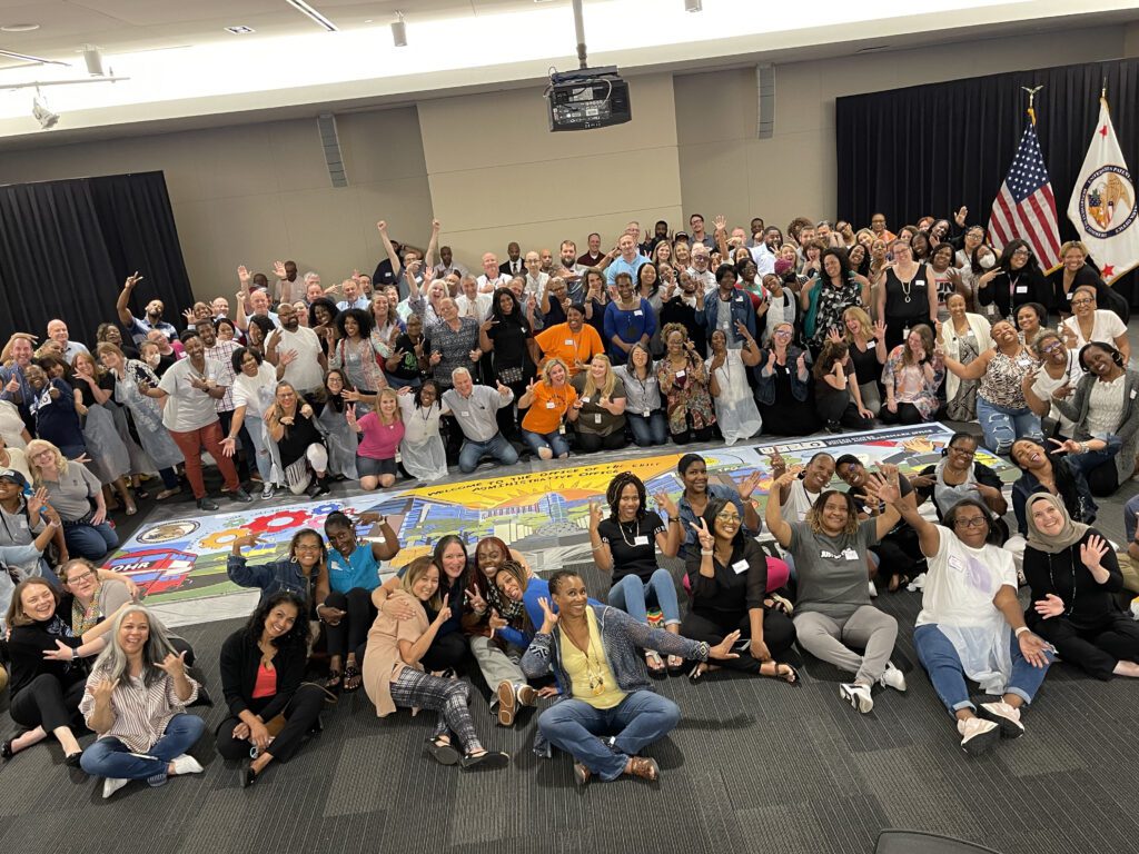 A large group of participants celebrates completing a collaborative mural during The Big Picture team building event, showcasing teamwork and creative achievement.