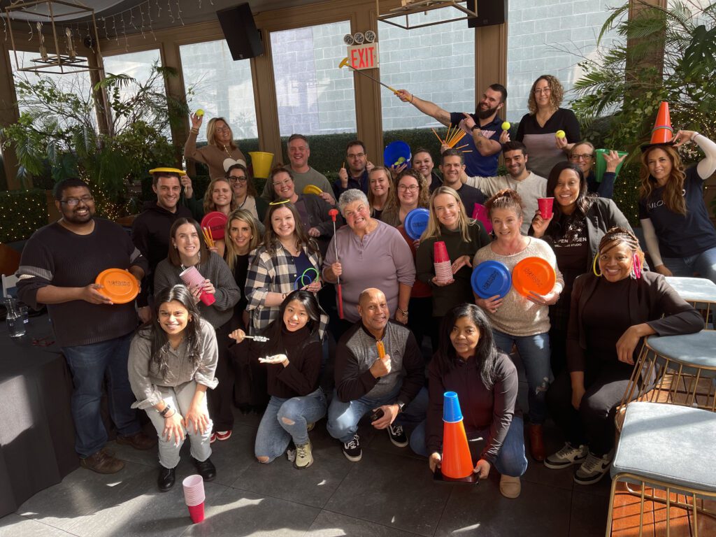 A large group of participants posing cheerfully with various props like cones, cups, frisbees, and rings, celebrating their participation in the In It to Win It team building event, showcasing a vibrant and fun team atmosphere.