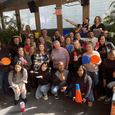 A large group of participants posing cheerfully with various props like cones, cups, frisbees, and rings, celebrating their participation in the In It to Win It team building event, showcasing a vibrant and fun team atmosphere.