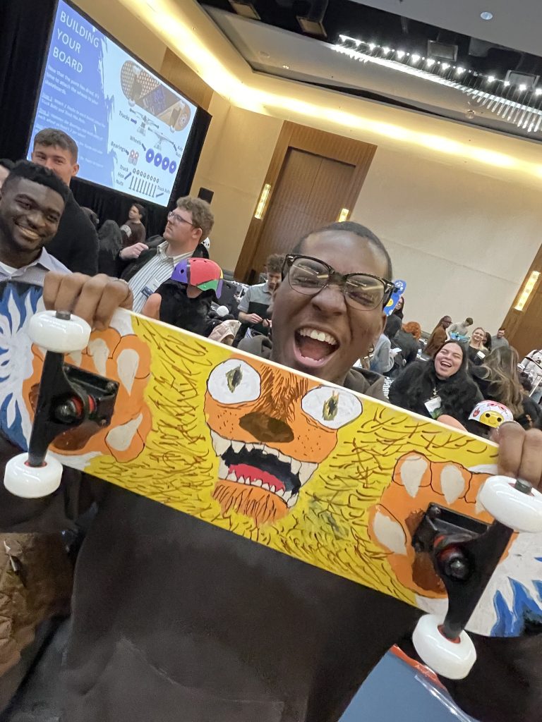 Excited participant proudly holds up a creatively designed skateboard during the Just Roll With It team building event, where teams build and decorate skateboards for charity.