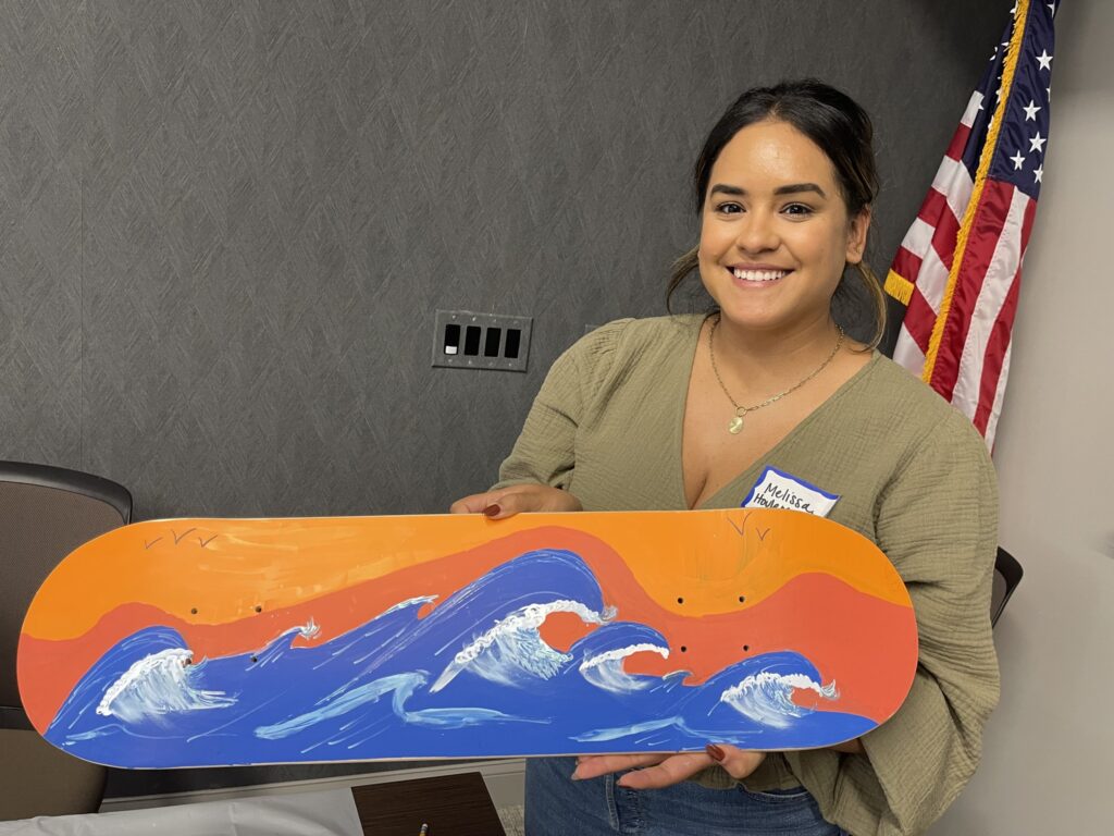 A smiling woman proudly holds up a skateboard that she painted during a team building event with her collages. The design features bright blue waves crashing against an orange and yellow sky, giving the impression of a sunset over the sea.