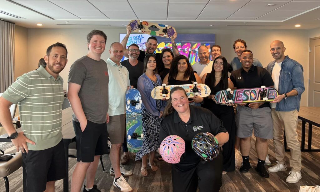 A group of smiling individuals stands together after completing a charitable skateboard build event. They proudly display decorated skateboards and helmets with colorful designs. One skateboard prominently says 'AWESOME' in bold letters, while helmets feature creative, abstract patterns. The group consists of men and women of various ages, all celebrating their accomplishments during this team building event.