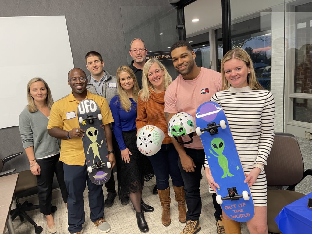 A group of eight people smiling and posing for a picture, holding custom-decorated skateboards and helmets. The skateboards and helmets feature fun designs, including aliens and 'UFO' themes. The group is participating in a team building event where they assemble and decorate skateboards to be donated to charity.