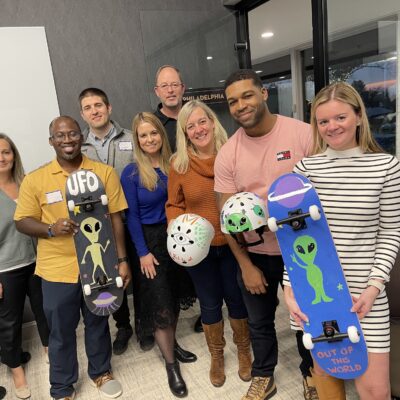 A group of eight people smiling and posing for a picture, holding custom-decorated skateboards and helmets. The skateboards and helmets feature fun designs, including aliens and 'UFO' themes. The group is participating in a team building event where they assemble and decorate skateboards to be donated to charity.