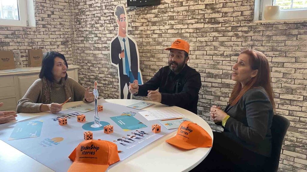 Participants engage in a Leadership Stories game, discussing and making leadership decisions, while wearing branded hats and working together at a table.