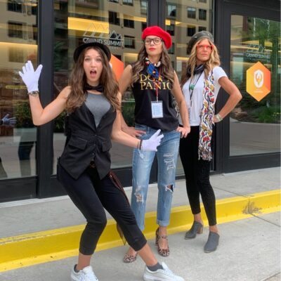 Three women dressed in creative costumes, including hats and gloves, posing energetically outside a venue during a Corporate Film Festival team building event, showcasing their excitement and team spirit.