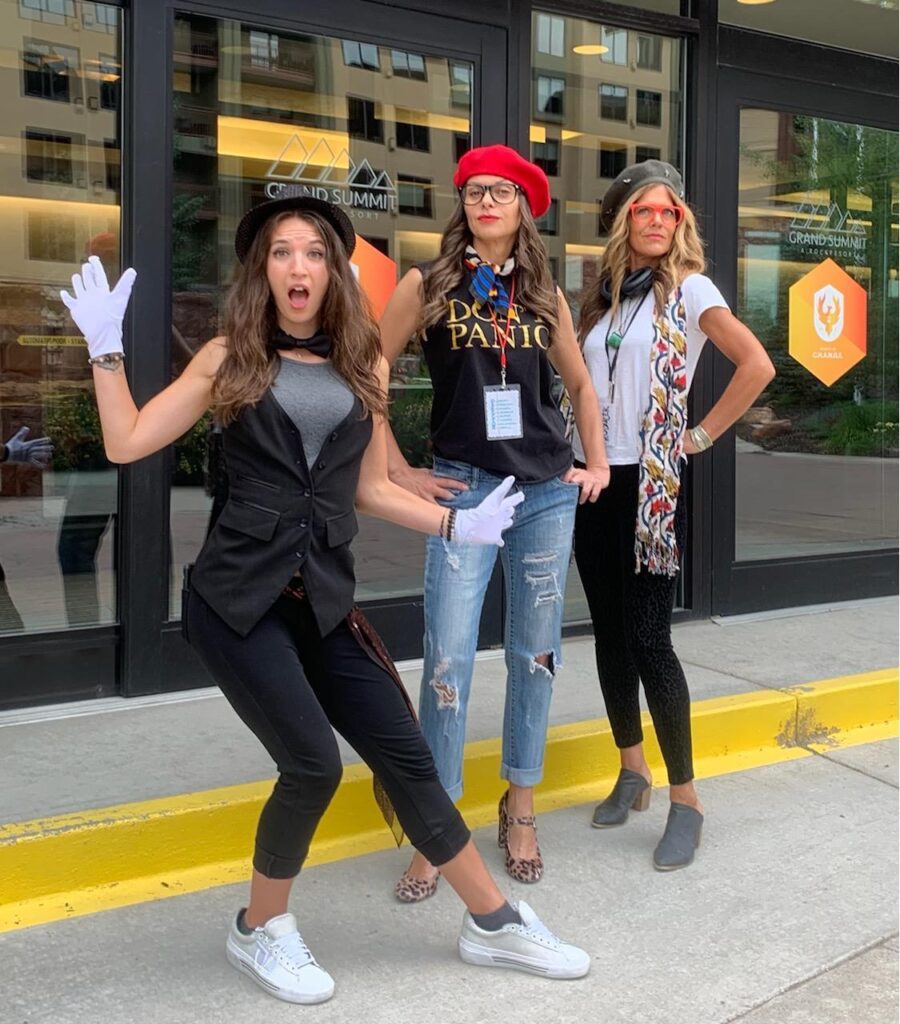 Three women dressed in creative costumes, including hats and gloves, posing energetically outside a venue during a Corporate Film Festival team building event, showcasing their excitement and team spirit.