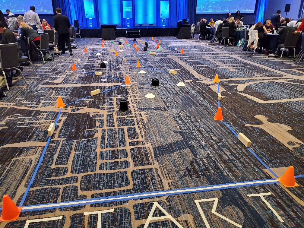 A robot obstacle course is set up indoors with orange cones, wooden blocks, and taped lines marking the track. Participants are preparing for a robot race, with 