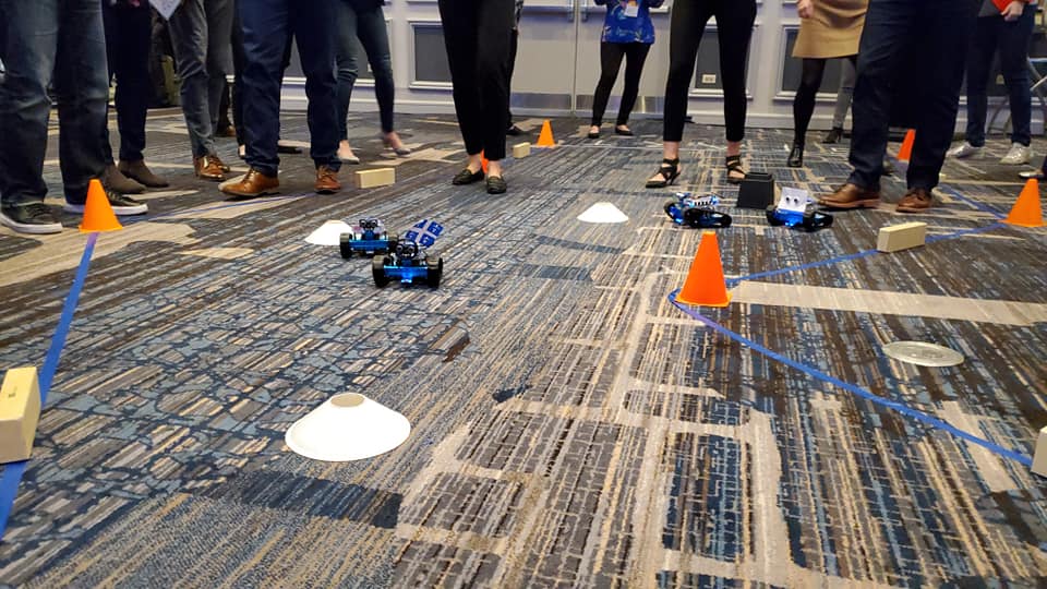 Participants are gathered around a designated race track, watching small robots they built navigate through an obstacle course marked by cones and barriers. The atmosphere is lively as teams cheer on their robots, showcasing the culmination of their team building activity.