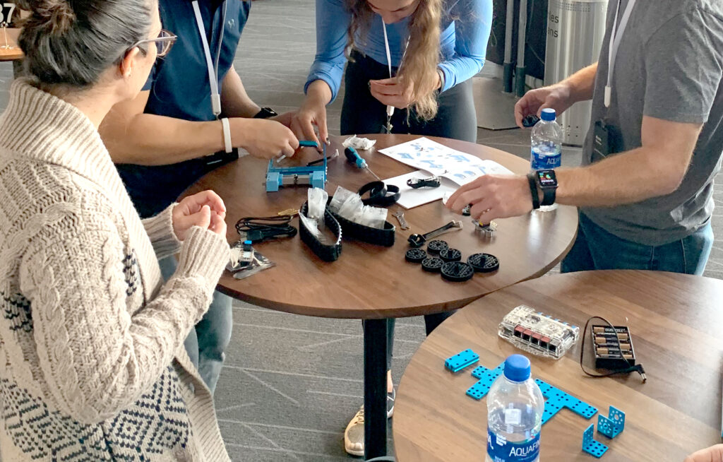 A group of people collaborate on building a robot at a table, using various robot parts, instructions, and tools during a robotics team building event. Bottled water and a battery kit are also on the table.