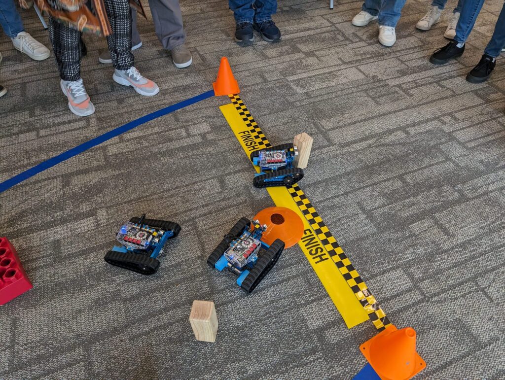 Three small robots are racing across a finish line in a friendly competition. Participants are gathered around, watching the robots as they navigate obstacles on the course. This image represents the final stage of a robotics team building event.