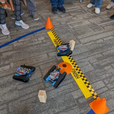 Three small robots are racing across a finish line in a friendly competition. Participants are gathered around, watching the robots as they navigate obstacles on the course. This image represents the final stage of a robotics team building event.