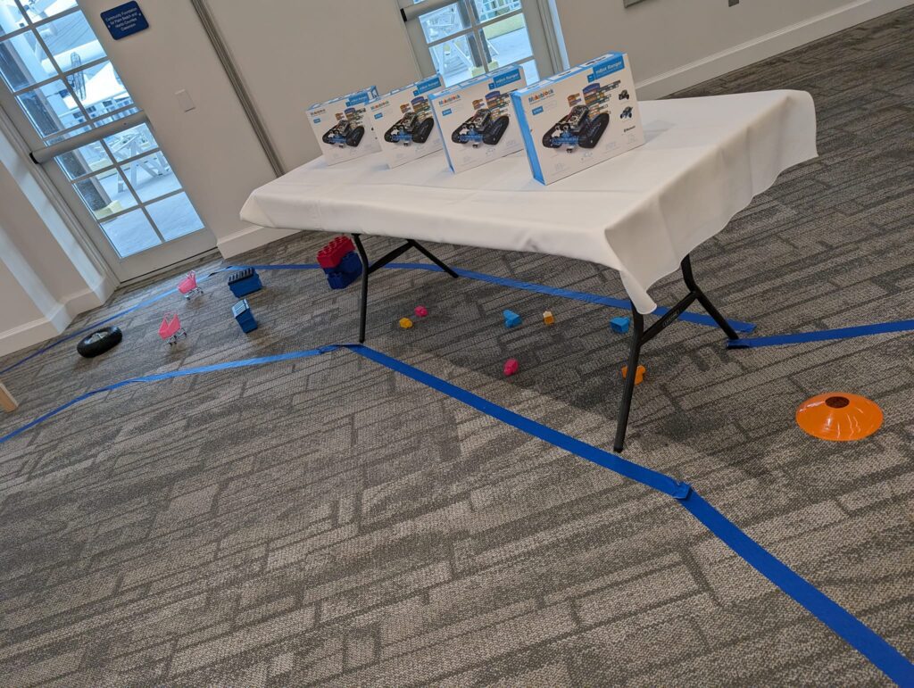 A table is set up with robot kits ready for assembly, placed in a room where a robotics team building event is taking place. Blue tape marks the floor for an obstacle course, alongside cones and other game objects.