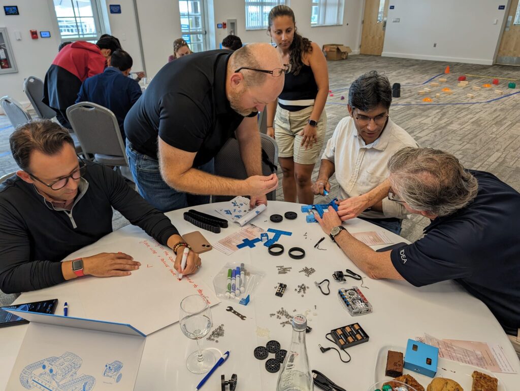 A group of participants is working together to assemble a robot. The table is filled with parts, tools, and instructions as they collaborate during a robotics team building event.