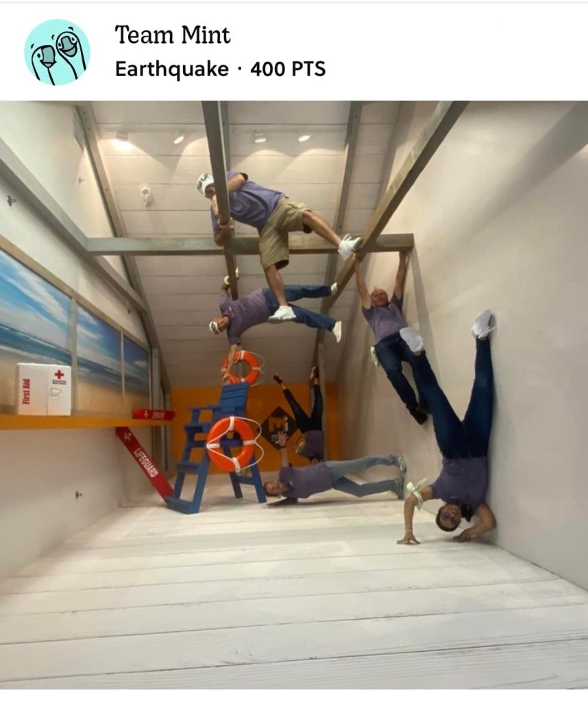 Team Mint posing creatively in an upside-down earthquake exhibit during a museum scavenger hunt. This team building activity promotes problem-solving, collaboration, and interactive fun in a unique setting.