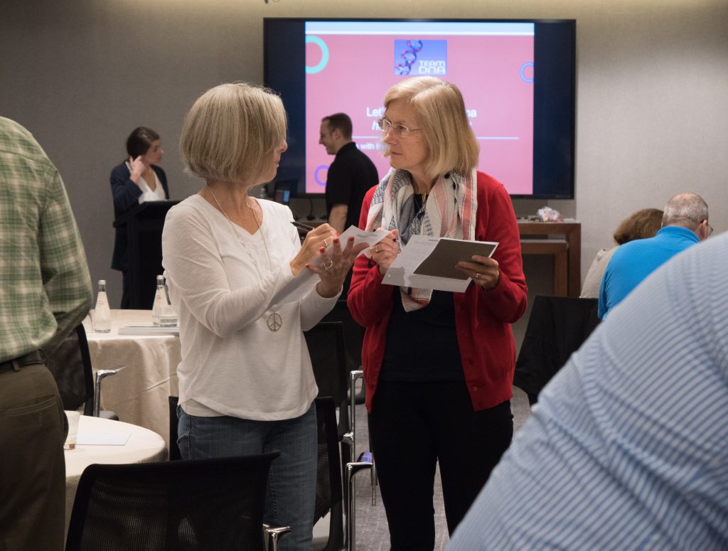 Two participants discussing and exchanging ideas during a Team DNA or Leadership DNA session, with other attendees engaged in the background.