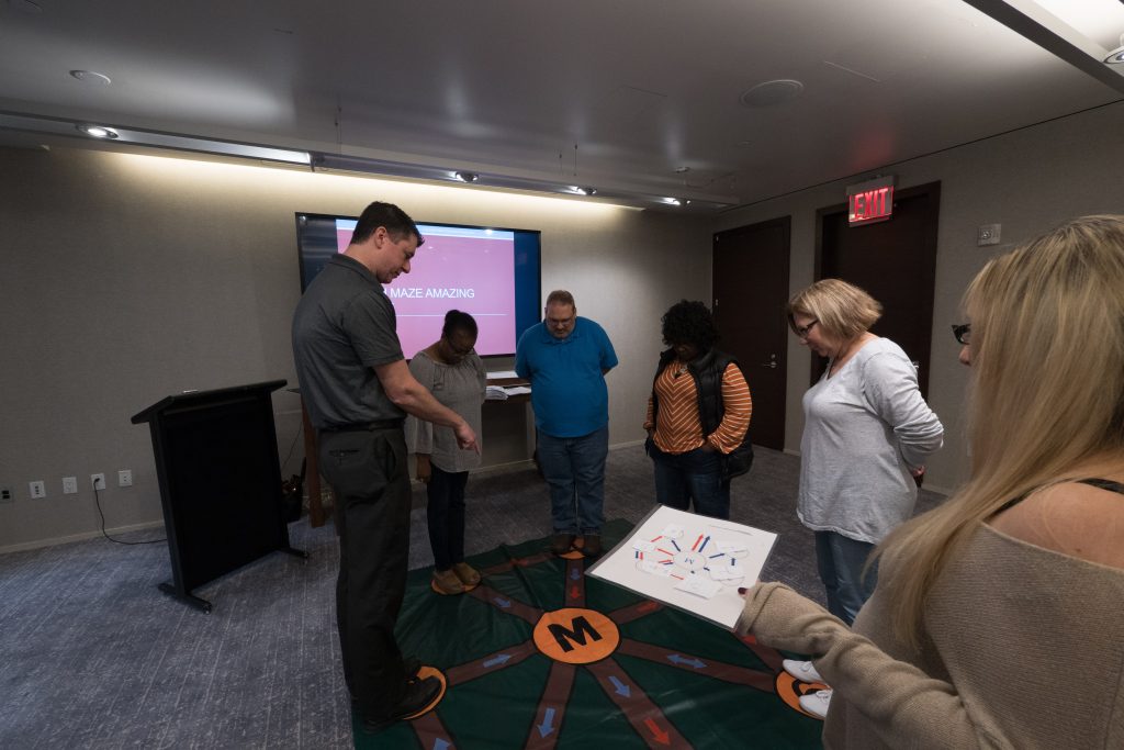 Participants engaging in a Team DNA activity, working together to solve a challenge while a facilitator leads the session with a visual display in the background.