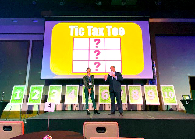 Two hosts stand on stage during the Play it Forward game show event, with a large screen behind them displaying the title 'Tic Tax Toe.' The event is a charitable team building activity where participants compete in pricing and trivia games to win for charity.