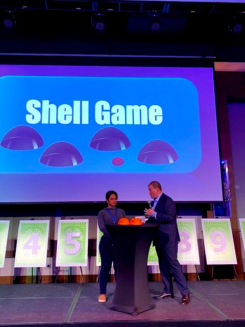 A contestant and host stand on stage during the Play it Forward charity game show, with the large screen behind them displaying the title 'Shell Game.' The contestant participates in a game where they must guess under which shell an item is hidden. Numbered boards are visible on the stage, representing different challenges in this fun, charitable team building event.