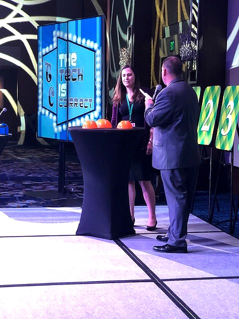 A host engages a contestant in the Play it Forward game show event on stage, with a screen in the background displaying the phrase 'The Tech is Correct.' The stage is set for a trivia challenge, and the contestant stands next to a table with orange buttons used in the game. Numbered boards are visible on the side, representing various challenges in this charitable team building activity.