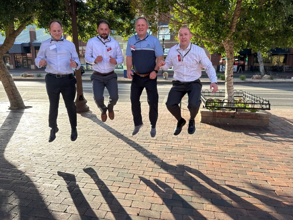 Four men participating in a team building activity captured mid-air while jumping on a sunlit brick pathway with trees and buildings in the background.