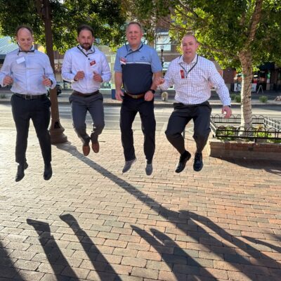 Four men participating in a team building activity captured mid-air while jumping on a sunlit brick pathway with trees and buildings in the background.