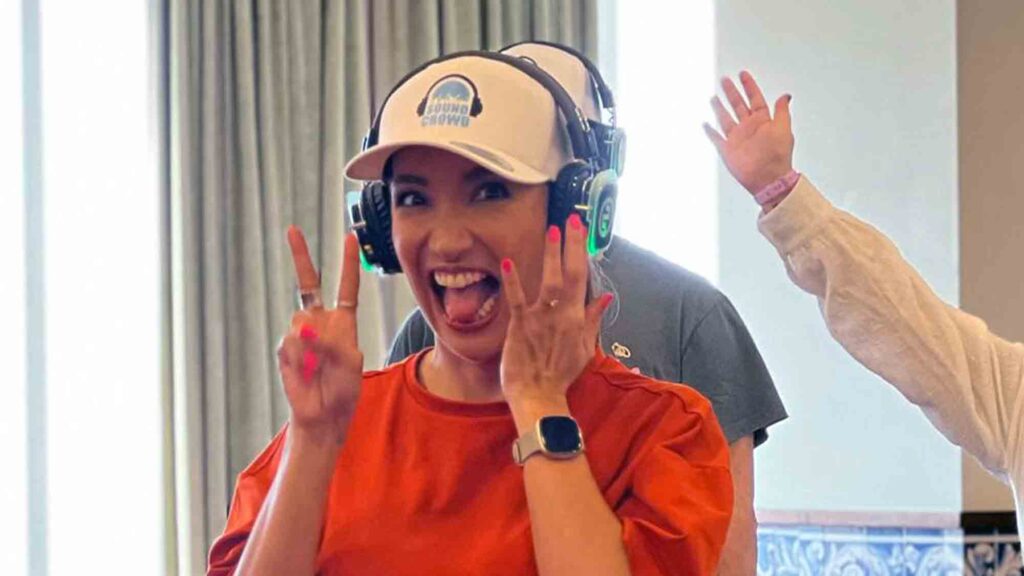 A woman wearing noise-cancelling headphones smiles widely, flashing two peace signs with her hands. She participates in a Sound Crowd team building event, where participants connect through sound and movement rather than verbal communication, fostering enthusiasm and group bonding.