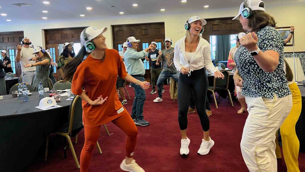 Participants in a Sound Crowd team building event laugh and dance while wearing noise-cancelling headphones. The group enthusiastically engages in the activity, which encourages non-verbal interaction through sound and movement, fostering fun and team connection.