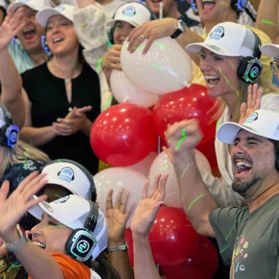 A large group of Sound Crowd participants cheering and smiling while wearing white caps and headphones. They are enthusiastically raising their hands, and red and white balloons are visible in the background, adding to the festive and vibrant atmosphere of the team building event.