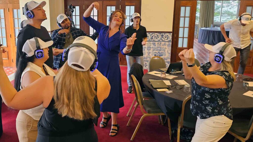 Participants at the Sound Crowd team building event dance and groove while wearing white caps and blue headphones. One woman in a royal blue dress is leading the group with expressive dance movements, while others follow along, all enjoying the music on their headsets.