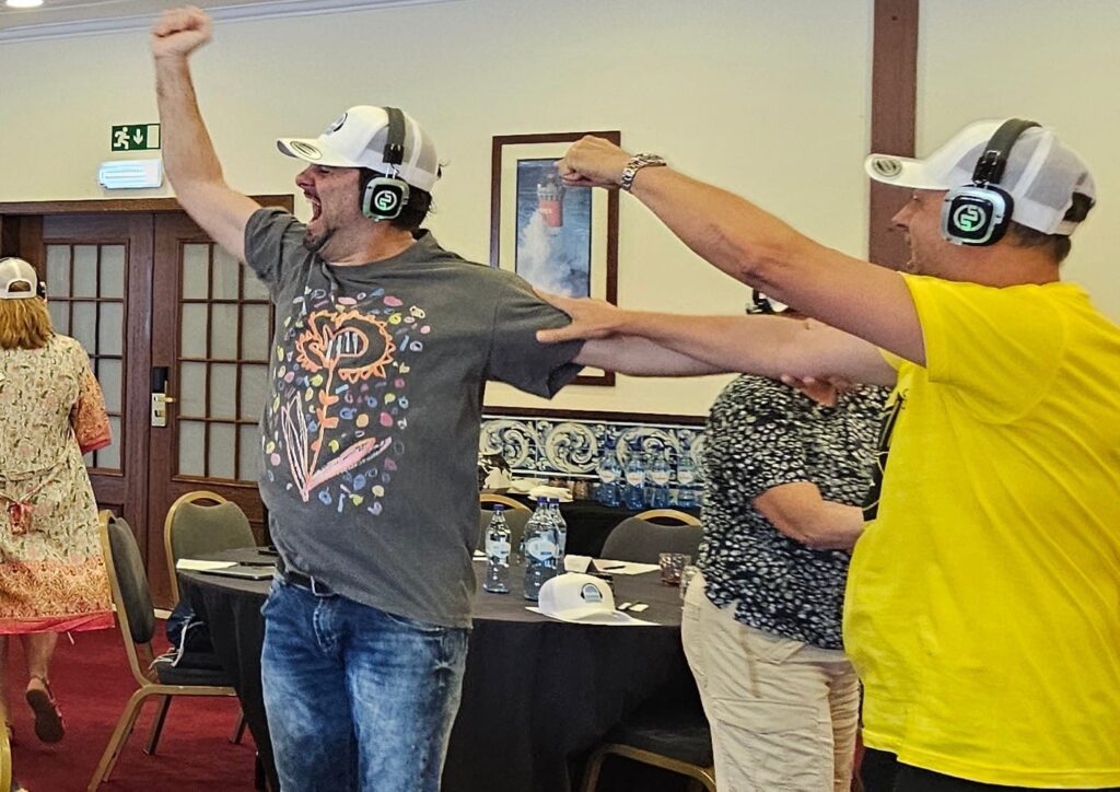 Two participants in a Sound Crowd team building event enthusiastically dance while wearing white caps and headphones. One man in a graphic t-shirt and jeans throws his arms up in excitement, while the other, wearing a yellow t-shirt, joins in with equally energetic movements.