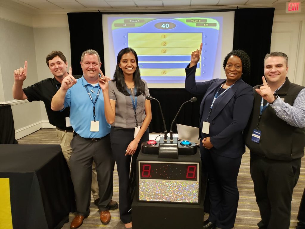 A team of five participants posing with enthusiasm and pointing to the screen, showing a game show setup during the Survey Says team building activity, highlighting team spirit, competition, and fun.