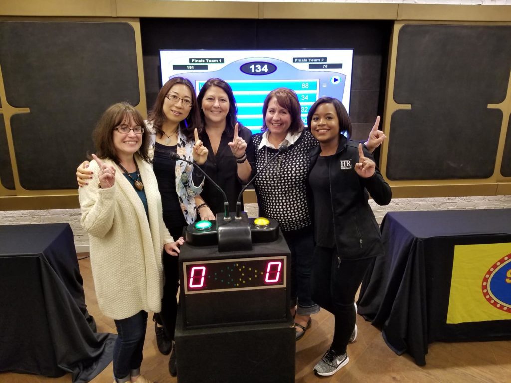 Five participants smiling and holding up their index fingers in front of a game show podium, celebrating their involvement in the Survey Says team building activity, with a game board displayed in the background.