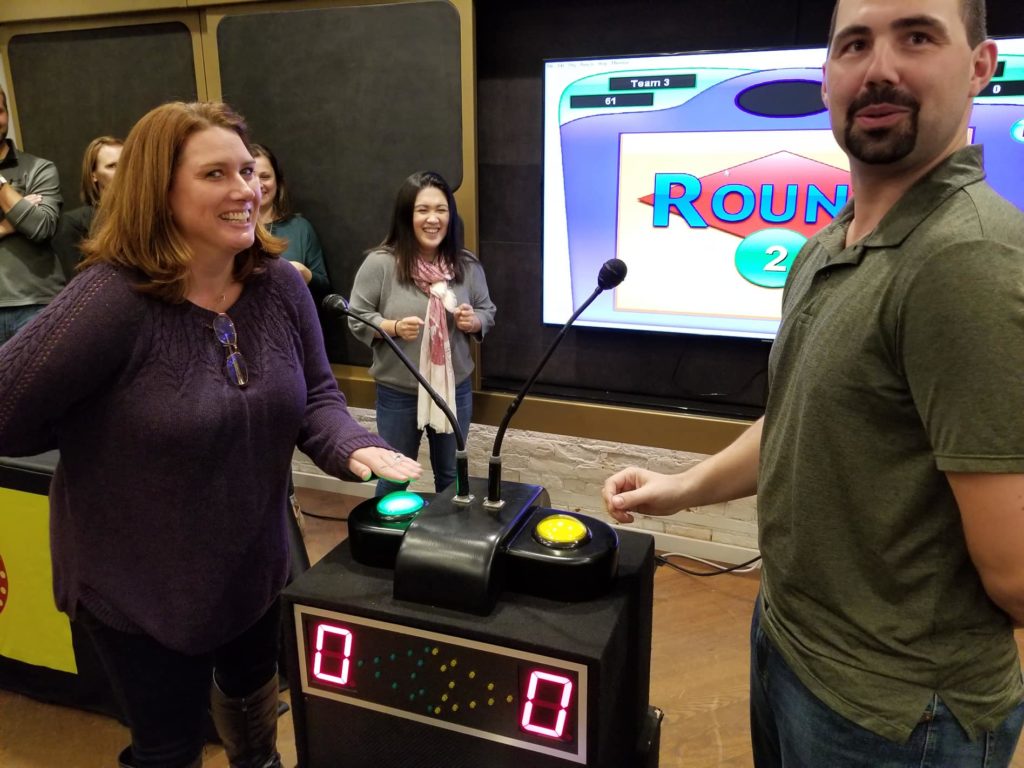 Two participants standing at a game show podium with buzzers, smiling and preparing to answer during the Survey Says team building event, with other participants watching in the background.