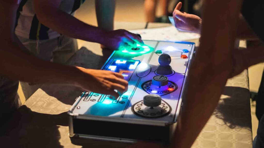 Team members collaborate on a brightly lit retro gaming console, using buttons, joysticks, and sliders during the Team Synergy team building event.