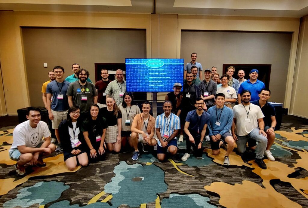 A large group of participants pose together in front of a Team Synergy retro gaming screen after a successful team building event, celebrating collaboration and teamwork.