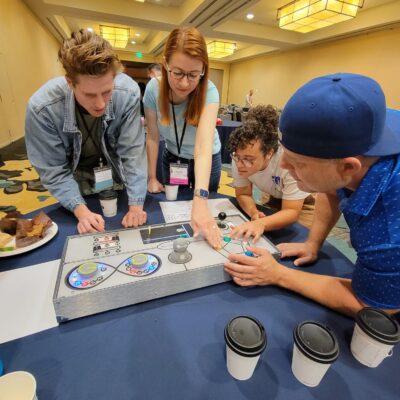 A group of participants works together on a Team Synergy gaming console, strategizing and solving challenges during a team building event in a conference room.