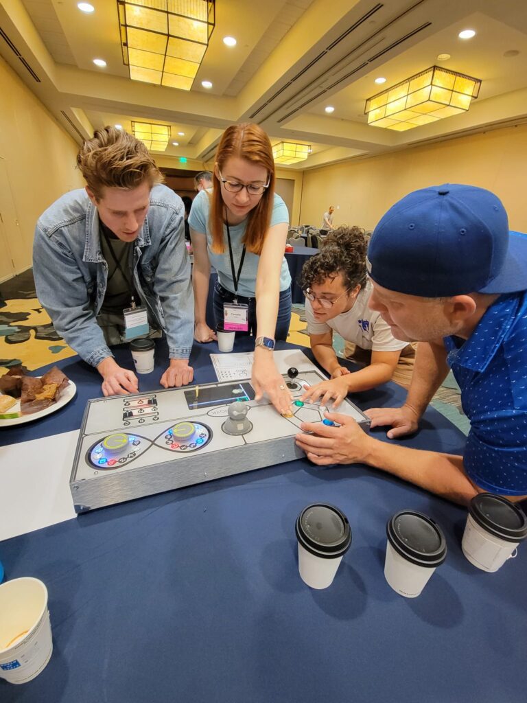 A group of participants works together on a Team Synergy gaming console, strategizing and solving challenges during a team building event in a conference room.